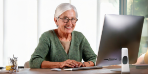 Seniorin sitzt glücklich an einem Schreibtisch vor einem dem Betrachter abgewandten Computerbildschirm
