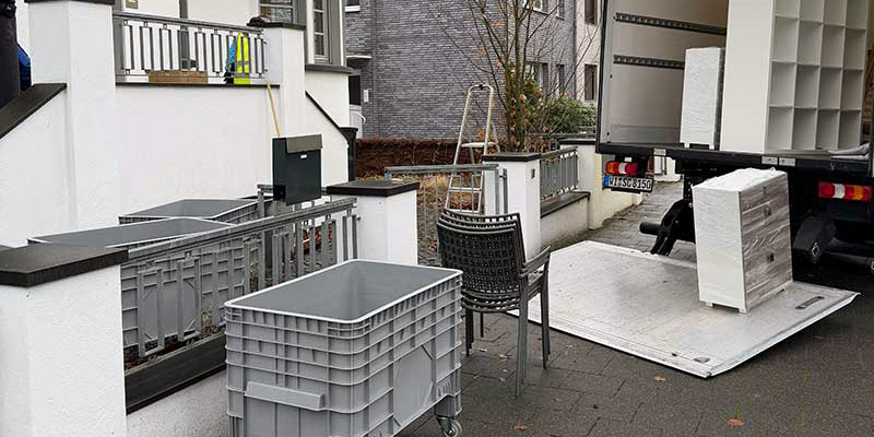 Vor einem weißen Wohnhaus steht ein Lkw mit offener Ladebordwand, daneben Rollcontainer und Kunststoffkisten auf dem Gehweg.